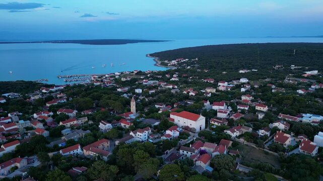 A breathtaking evening aerial drone panorama of old village on Silba Island, Croatia. The shot showcases the vibrant turquoise harbor, a docked ferry, and the charming coastal village stretching