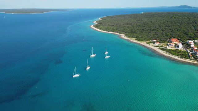 An aerial panoramic view of the tranquil turquoise bay on Silba Island, Croatia, featuring anchored sailboats along a lush, green Mediterranean coastline with charming red-roofed village houses under