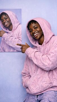 Joyful Black woman in a pink hoodie laughing and pointing at a portrait