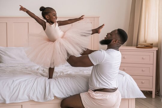 Excited black father and daughter playing together and dancing on a large bed in a bedroom