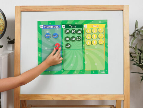 Close-up of a child's hand using colorful number manipulatives on a place value chart with hundreds, tens and ones during a hands-on mathematics learning activity.
