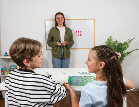Female teacher stands by a whiteboard with an addition problem while a boy and girl sit at a table using place value math manipulatives during an elementary mathematics lesson.