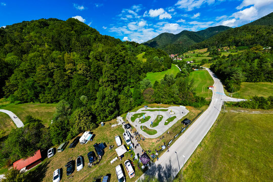 A wide aerial view of the asphalt pumptrack park in Zagorje ob Savi, Slovenia, surrounded by green fields, rolling hills, a railway line, and a bright blue sky filled with fluffy white clouds.