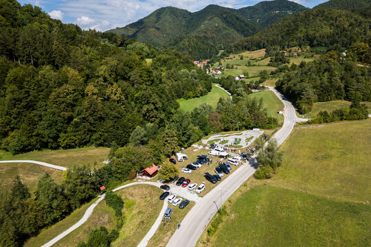 A wide aerial view of the asphalt pumptrack park in Zagorje ob Savi, Slovenia, surrounded by green fields, rolling hills, a railway line, and a bright blue sky filled with fluffy white clouds.