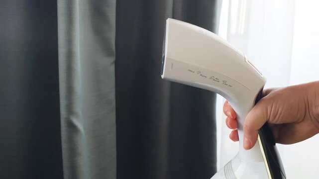 Vertical steam cleaning process on window curtains using handheld steamer. Effective steam cleaning smooths gray drapes, eliminates creases, and freshens home textiles in modern room setting.