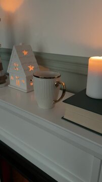 Vertical B-roll close up shot of female hand putting coffee mug on white fireplace mantel with lit candle and glowing house decor. Cozy hamptons and hygge living room lifestyle concept. 4K video.