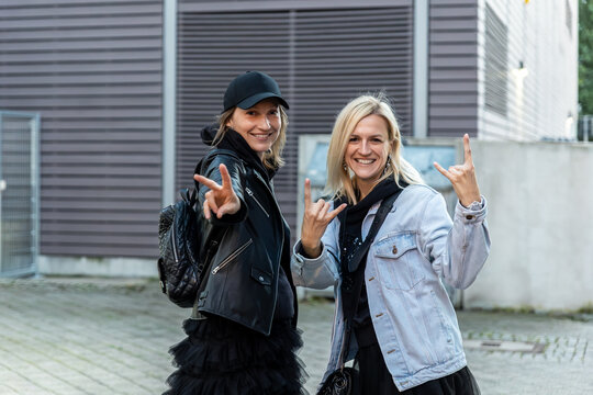 Two stylish young adult women middle aged showing rock and peace hand signs in casual street fashion. Friendly portrait captures confidence, friendship, music, urban style, and playful energy