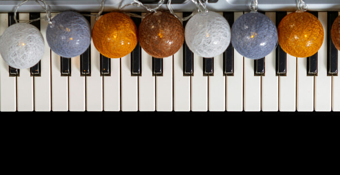 Decorative cotton ball string lights rest on top of black and white piano keys. A colorful garland of round bulbs creates a festive pattern across the surface. Winter holiday music and celebration