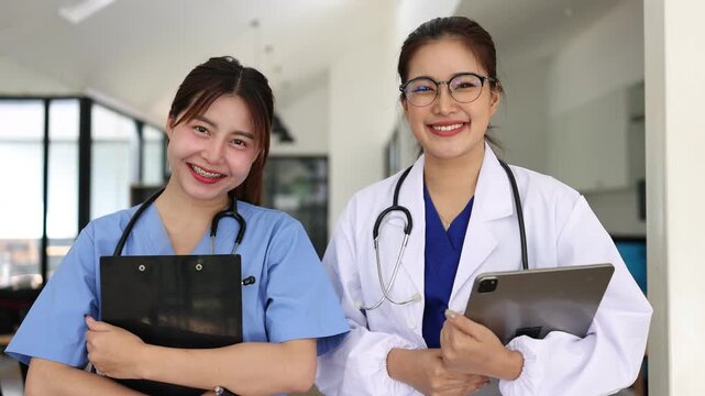 Portrait of two confident and smiling Asian female medical professionals, a doctor and a nurse, holding a clipboard and a digital tablet in a hospital hallway.