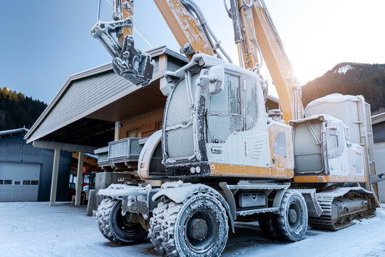 Scenic view frozen hydraulic excavators standing outdoors winter conditions covered snow ice industrial building alpine area. Cold weather impacts machinery operation hydraulic systems reliability