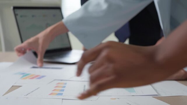 Multiple hands gesture toward data on a tablet screen, printed graphs and documents covering the table in a busy strategy meeting.