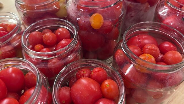Multiple clear glass jars filled with red and orange cherry tomatoes, some jars containing liquid and others packed with whole vegetables on a light surface