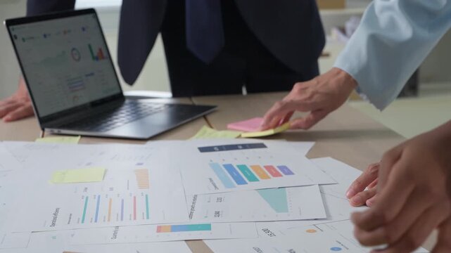 Multiple hands gesture toward data on a tablet screen, printed graphs and documents covering the table in a busy strategy meeting.
