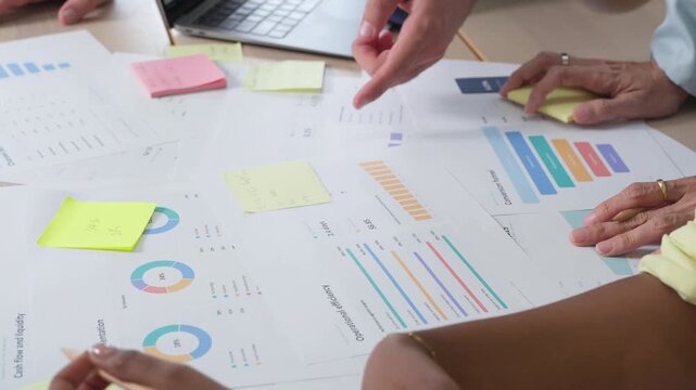 A detailed view of hands on a laptop and paperwork, colleagues collaborating over data charts in a fast-paced office meeting.