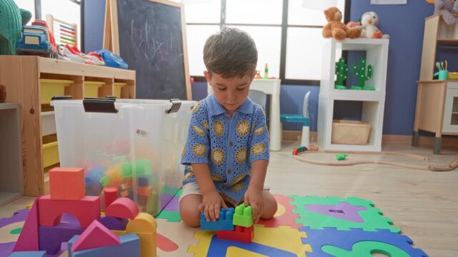 Boy stacking colorful plastic blocks with bare hand on foam alphabet mat inside a building playroom; focused concentration.