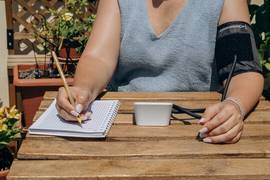 Woman Checking Blood Pressure At Home Outdoors While Recording Readings In Notebook On Wooden Table