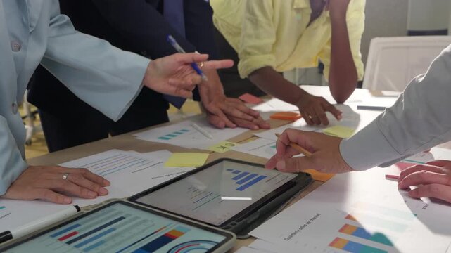 Multiple hands gesture toward data on a tablet screen, printed graphs and documents covering the table in a busy strategy meeting.