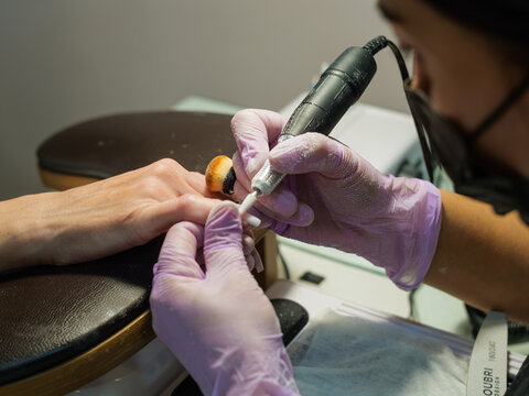 A horizontal view of a master in a black apron and purple gloves conducting a hardware manicure procedure on a client's hand in a salon.