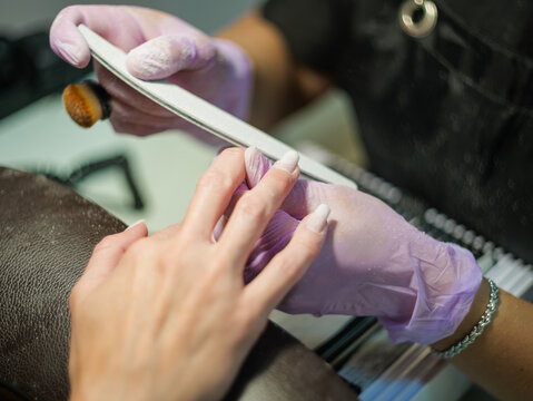 A horizontal close-up shot of a manicurist in purple gloves filing and shaping a client's fingernails using a nail file in a salon.