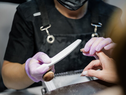 A horizontal close-up view of a beautician using a nail file to shape and smooth a client's fingernails at a beauty salon workstation.   