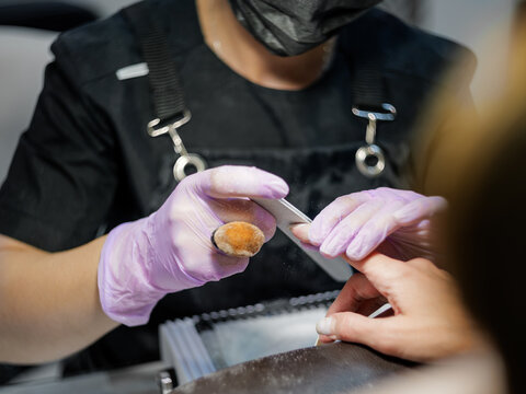 A horizontal photograph showing a professional manicurist filing the nail plate on a client's hand in a modern nail salon.   