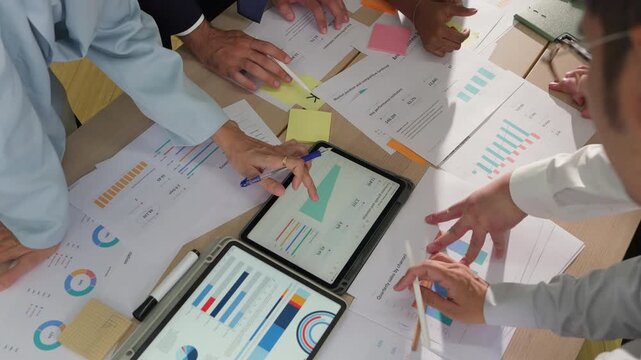 Overhead view of multiple hands pointing at financial charts on a tablet, surrounded by printed reports and colorful data visualizations on a busy desk.