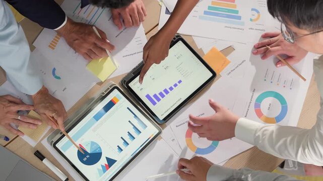 Overhead view of multiple hands pointing at financial charts on a tablet, surrounded by printed reports and colorful data visualizations on a busy desk.