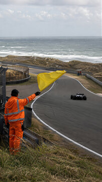 Race marshal waves a yellow flag beside a single-seater car on a coastal circuit. The marshal wears a bright orange safety suit. Motor racing competition under caution at a seaside track.