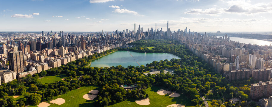 Aerial view displays Central Park reservoir surrounded by trees, baseball fields, and the Midtown Manhattan skyscraper cityscape under clear skies in New York City.