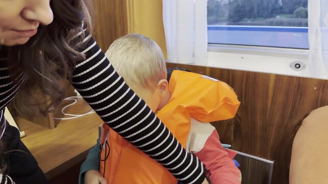 A mother adjusts a life vest on her child in a cruise ship cabin on the Neva River, Saint Petersburg. Her caring help makes the boy feel safe and secure. The moment brings reassuring comfort.