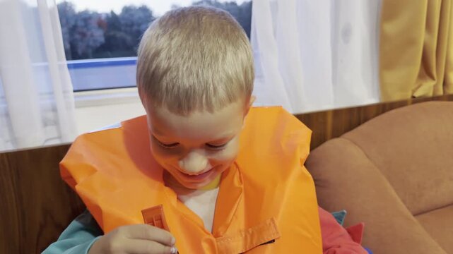 A happy boy smiles while wearing an orange life jacket inside a cruise ship cabin on the Neva River, Saint Petersburg. He feels excited and safe. The cozy room brings joyful family moments.