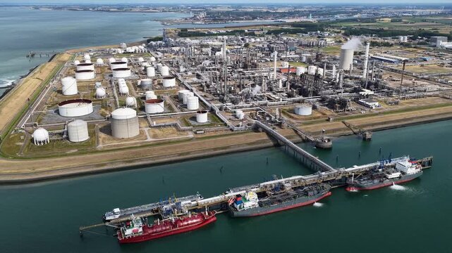 Aerial drone view of tanker ships docked at an industrial terminal, with extensive pipelines connecting vessels to large storage tanks and processing facilities along the waterfront.