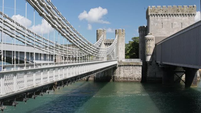 Historic iron suspension bridge designed by Thomas Telford beside stone castle walls and towers over green water under a bright summer sky