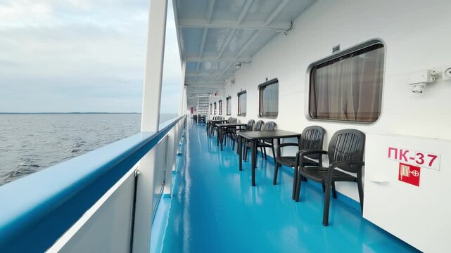 An empty cruise ship deck glides smoothly across Lake Ladoga. Tables and chairs rest along the blue promenade under a cloudy sky. The serene view brings a calm and peaceful feeling.