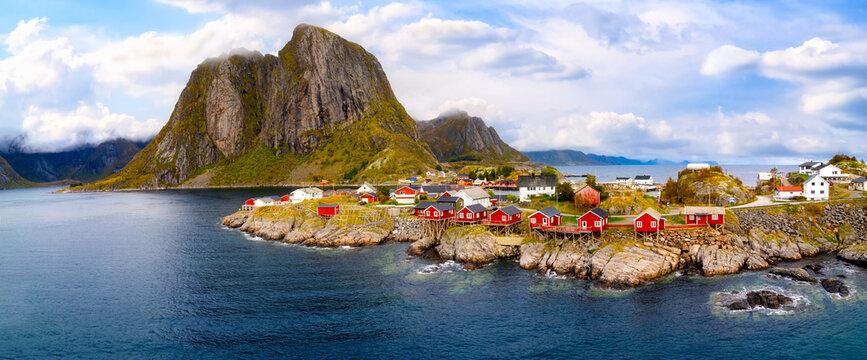 Autumn coastal scenery of Hamnoy in the Lofoten Islands with traditional red fishing houses, rocky shoreline, blue sea and dramatic mountains beneath a cloudy sky.