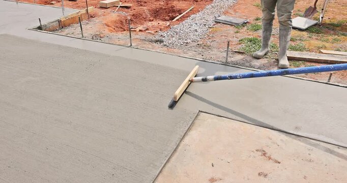 Worker uses long brush tool to smooth surface of wet concrete being poured at construction site.