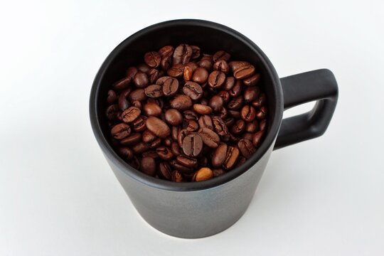 Mainz, Rhineland Palatinate, Germany: Roasted coffee beans filling black ceramic mug on clean white background.