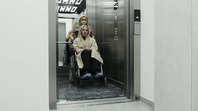Nurse pushes wheelchair with disabled patient out of elevator. Experienced doctor transports patient toward treatment room. Slow motion