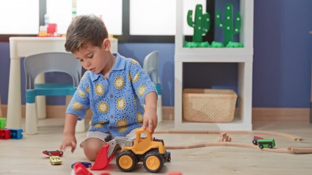 Boy kneeling pushes a yellow toy loader into a scoop amid cars and wooden tracks in a playroom; concentration.