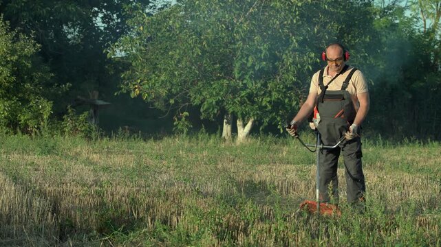 Landscaper using a string trimmer to cut dry grass and weeds in a sunny rural meadow surrounded by trees and natural vegetation
