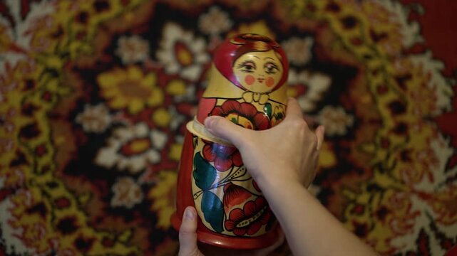 Woman's hands open a painted Russian matryoshka doll, revealing inner layers on a floral vintage rug.
