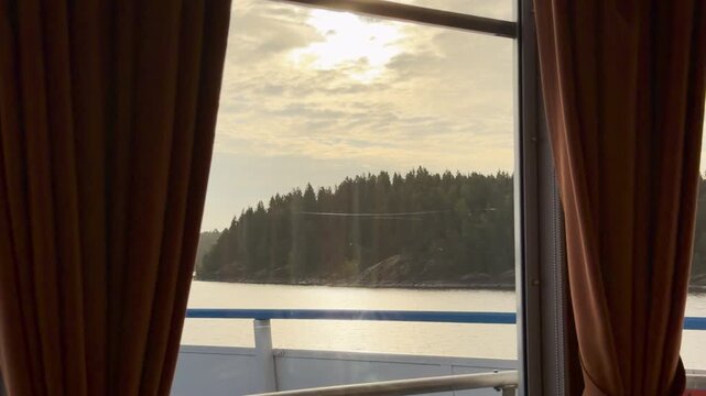 A cruise ship window frames a view of pine forest shores on Lake Ladoga. Warm sunlight shines over the calm water. The peaceful scenery brings a serene and cozy feeling.