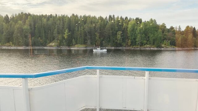 A cruise ship sails smoothly across Lake Ladoga near dense forests. A small boat rests on calm, dark waters under overcast skies. The serene landscape inspires a peaceful sense of wonder.