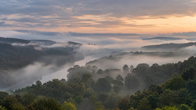 Morning mist rolling over scenic river valley surrounded by green forested hills at sunrise. Concept of fog, foggy sunrise, landscape. Copy space.