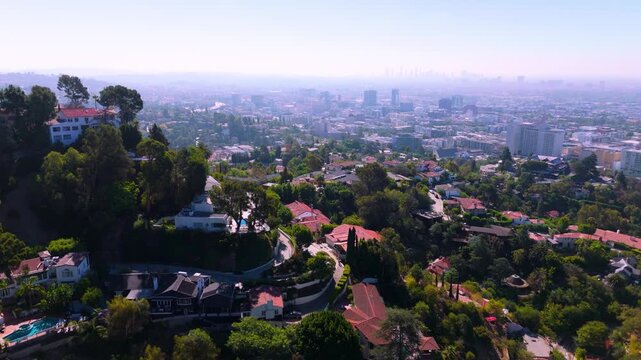 Aerial over Hollywood Hills luxury homes with red tile roofs swimming pools palm trees and winding hillside road. Inspiring drone view of hazy downtown Los Angeles skyline on sunny summer day.