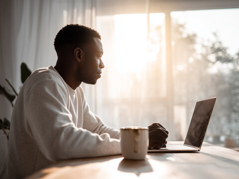African American man sitting indoors at desk working remotely on laptop with cup of coffee in morning
