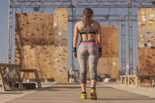 Female runner standing back to camera, preparing for obstacle course, sporty outfit, high-resolution, dynamic outdoor training scene, realistic textures, motivational sports concept.
