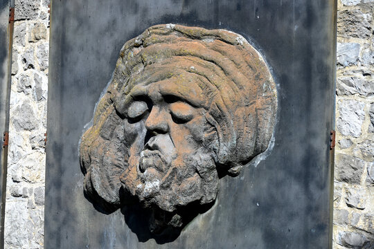 une t&ecirc;te sculpt&eacute;e sur une vieille plaque de m&eacute;tal avec des cheveux ou un turban, des gros yeux ferm&eacute;s, le nez et la bouche. Le visage est de couleur rouille sur un fond gris.