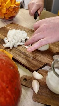 A person uses a knife to chop a white onion on a wooden cutting board, with bowls of chopped orange vegetables and garlic cloves nearby on a light wooden surface