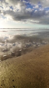 Calm sea waves washing over the wet sandy shore while the cloudy blue sky is reflecting on the water surface during a peaceful morning, showing the concept of tranquility and nature